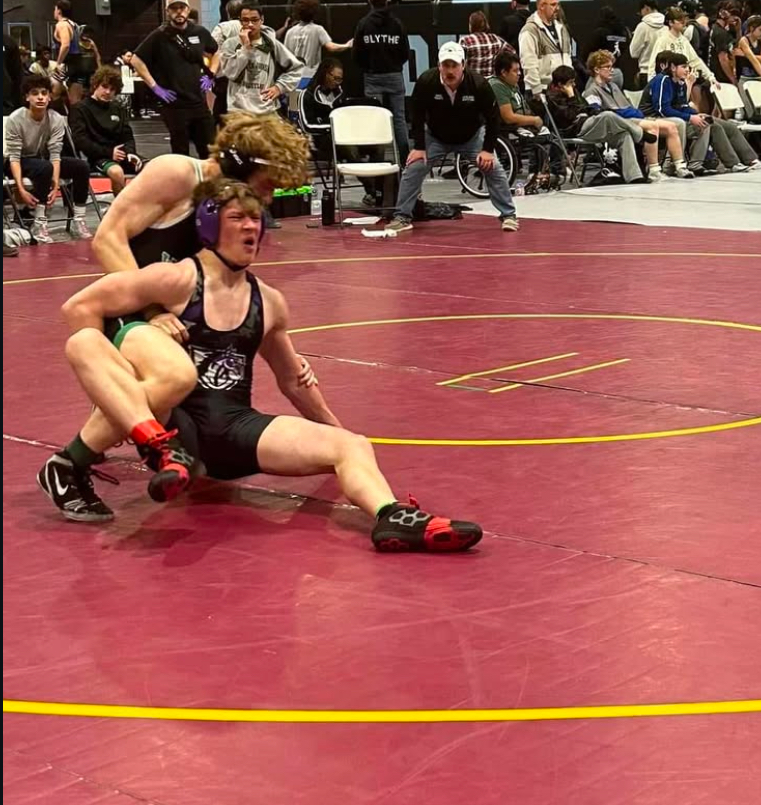 Junior Filippos Kakos leg riding his opponent and bringing him to the floor, while coach Brian Frantz watches closely in the background. (Courtesy WJ Athletics on Instagram)