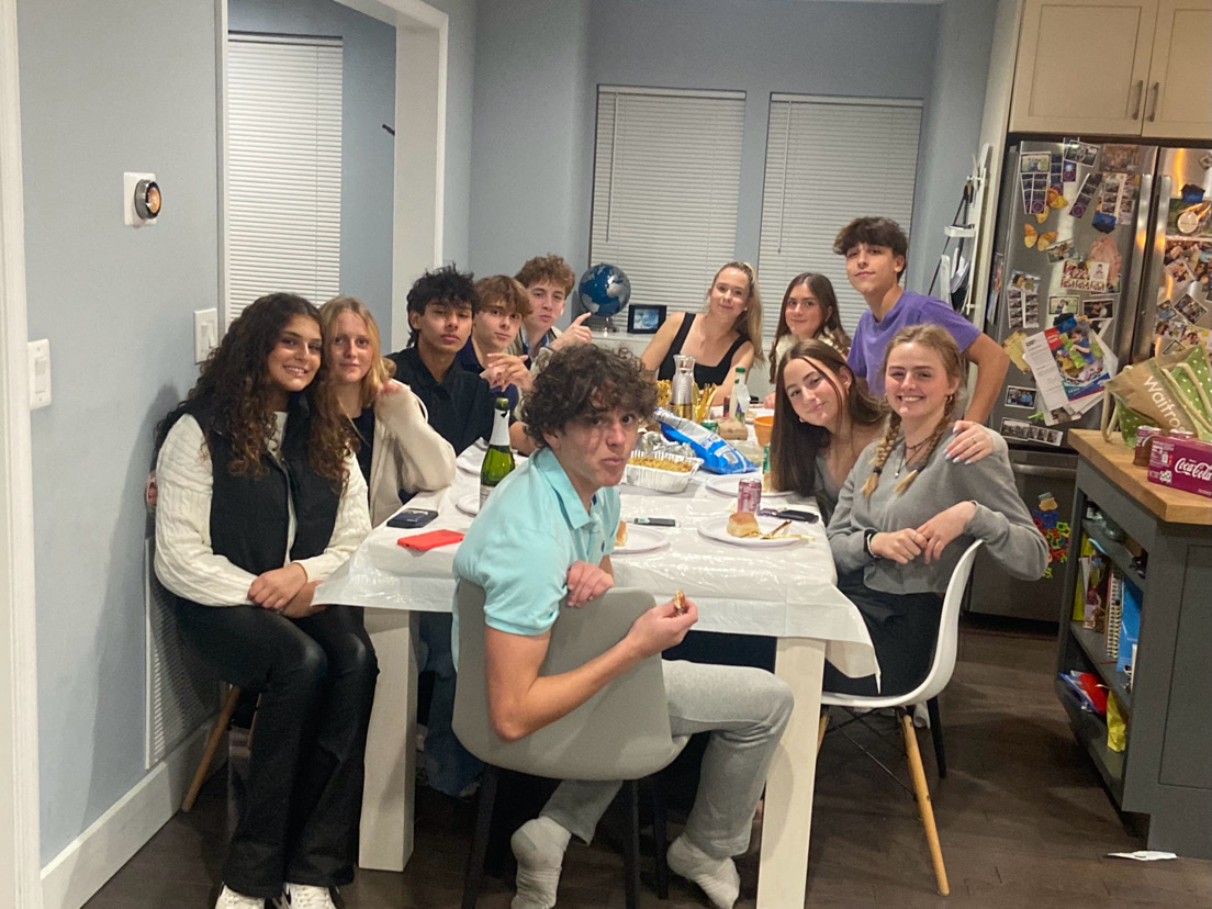 A friend group sits around the table, celebrating Friendsgiving. They did a potluck, where each person brought their own food, to elevate the holiday spirit.