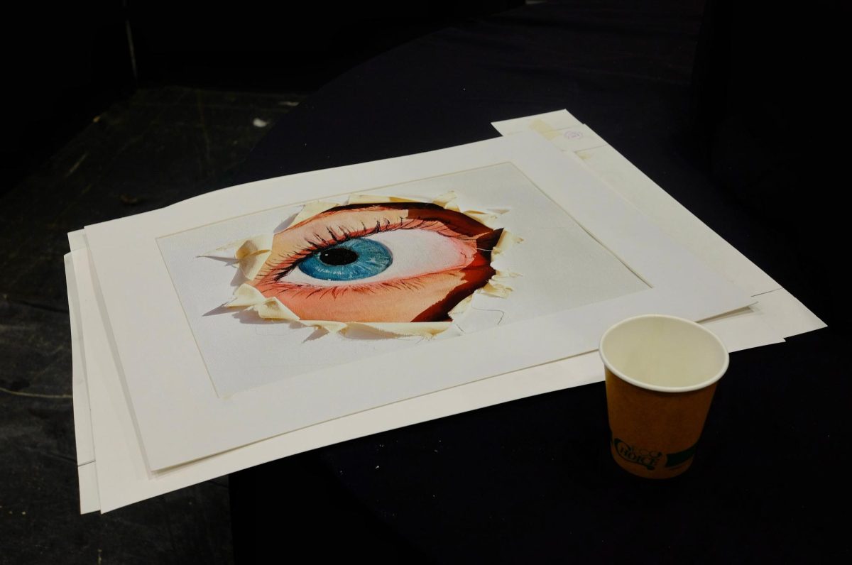 A paper cup filled with pins for mounting artwork sits beside a stunningly realistic depiction of a blue eye, created by junior Melin Öztürk of AP 2D Art and Design class. The piece uses a 3D torn paper effect, creating a striking sense of depth that catches the attention of each passerby.